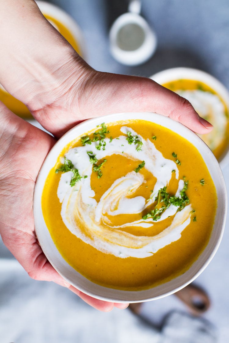 Hands holding a bowl with curry coconut carrot soup garnished with ginger cream and parsley.