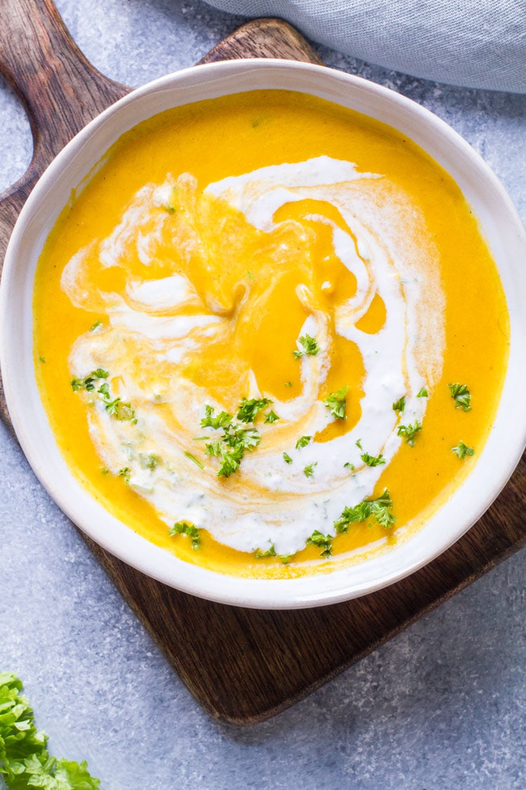 White bowl with carrot soup and swirls of ginger cream on a wooden cutting board.