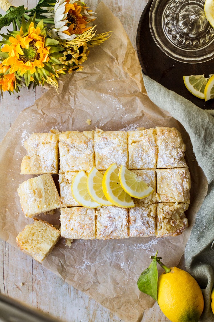 Lemon Cake with lemon slices and powdered sugar on top. Background is parchment paper and a green towel. Some yellow flowers and lemons in the corner.