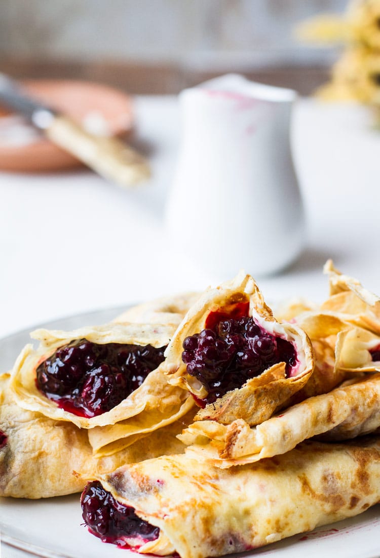 A stack of crepes filled with lemon cream cheese and blackberry jam. White plate, white background and white mug with blackberry jam.