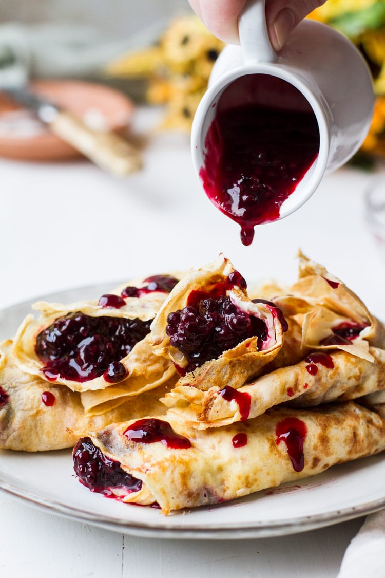 A stack of crepes filled with lemon cream cheese and blackberry jam. White plate, white background and white mug with blackberry jam pouring over the crepes.