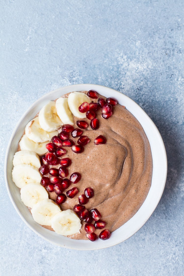 Bowl with chocolate chia pudding, decorated with banana slices and pomegranate to the left. Blue background, flatlay.