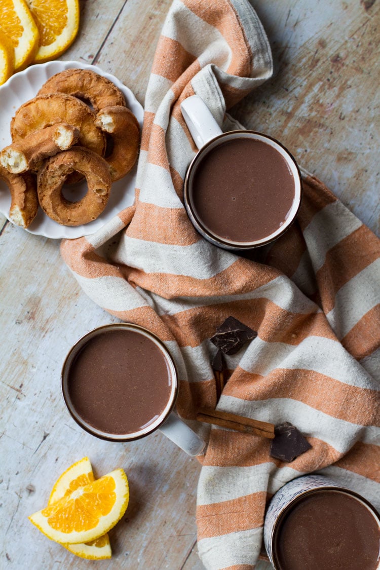 Two cups with hot chocolate, one orange striped towel between them and donuts on the side. Flatlay.