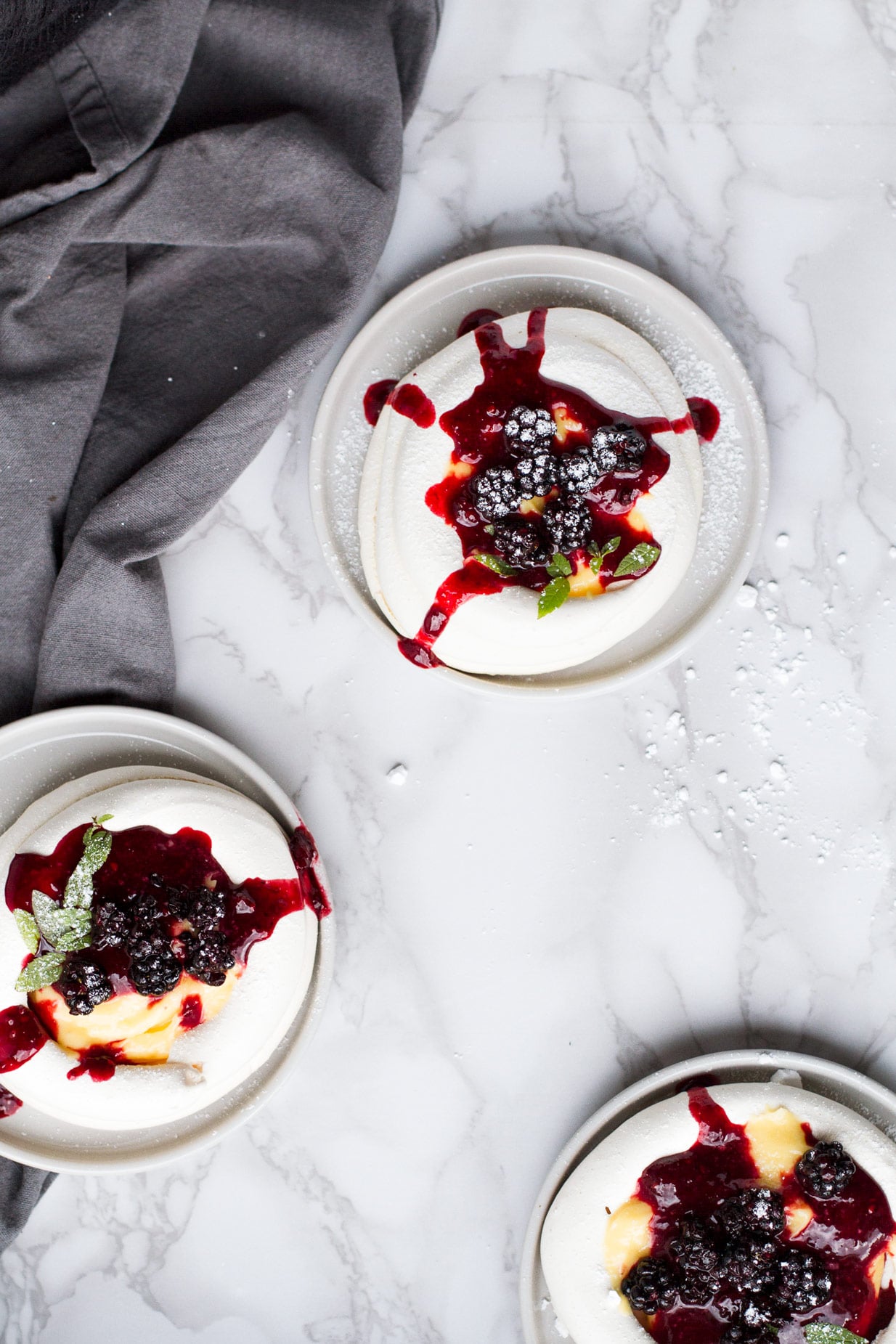 Three plates of pavlova nests with blackberry syrup running down their sides. Flatlay.