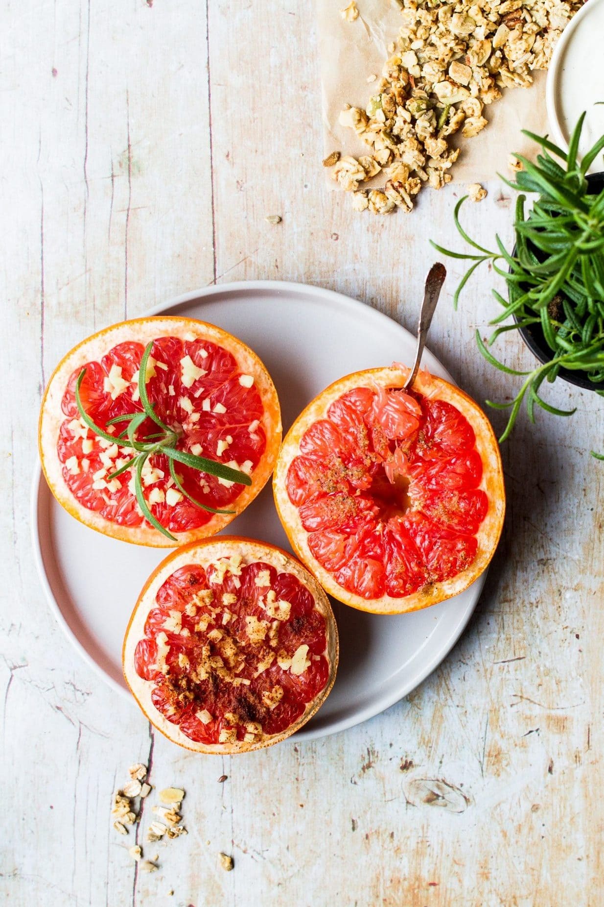 Bird's eye view of three half grapefruits on a beige plate and a light wooden background. One has a spoon in, one has rosemary on and one has spice sprinkles. A little rosemary and granola in the corner of the shot.
