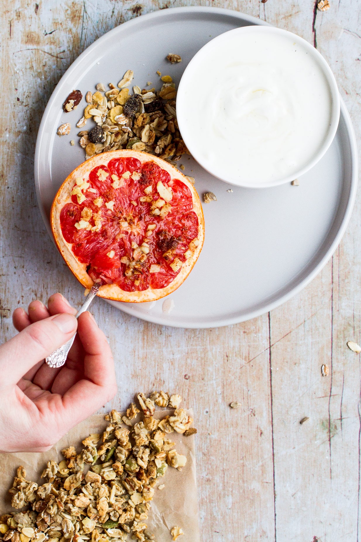 Birsd's eye view of a beige plate with one small bowl of yoghurt, a drizzle of granola and a baked grapefruit. A hand scooping up a piece with a spoon. Light wooden background