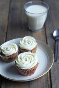 Three carrot cake cupcakes with floret shaped cream cheese frosting on a white plate. Milk in the background.