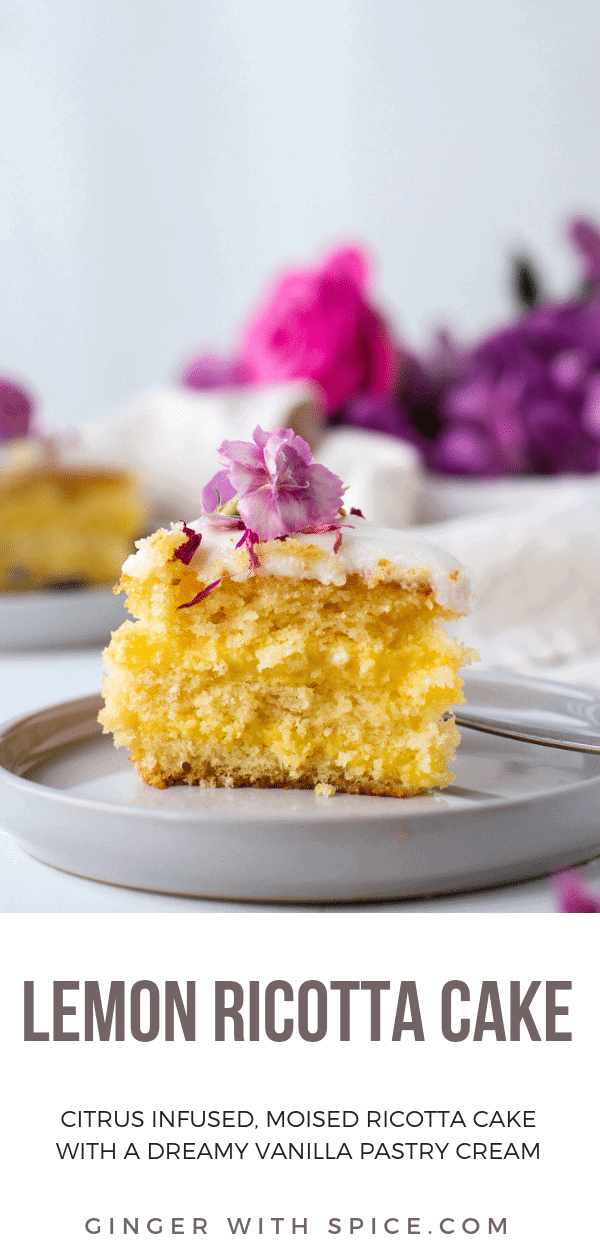 Yellow slice of cake on a grey plate with a purple flower on top. More purple flowers in the background.