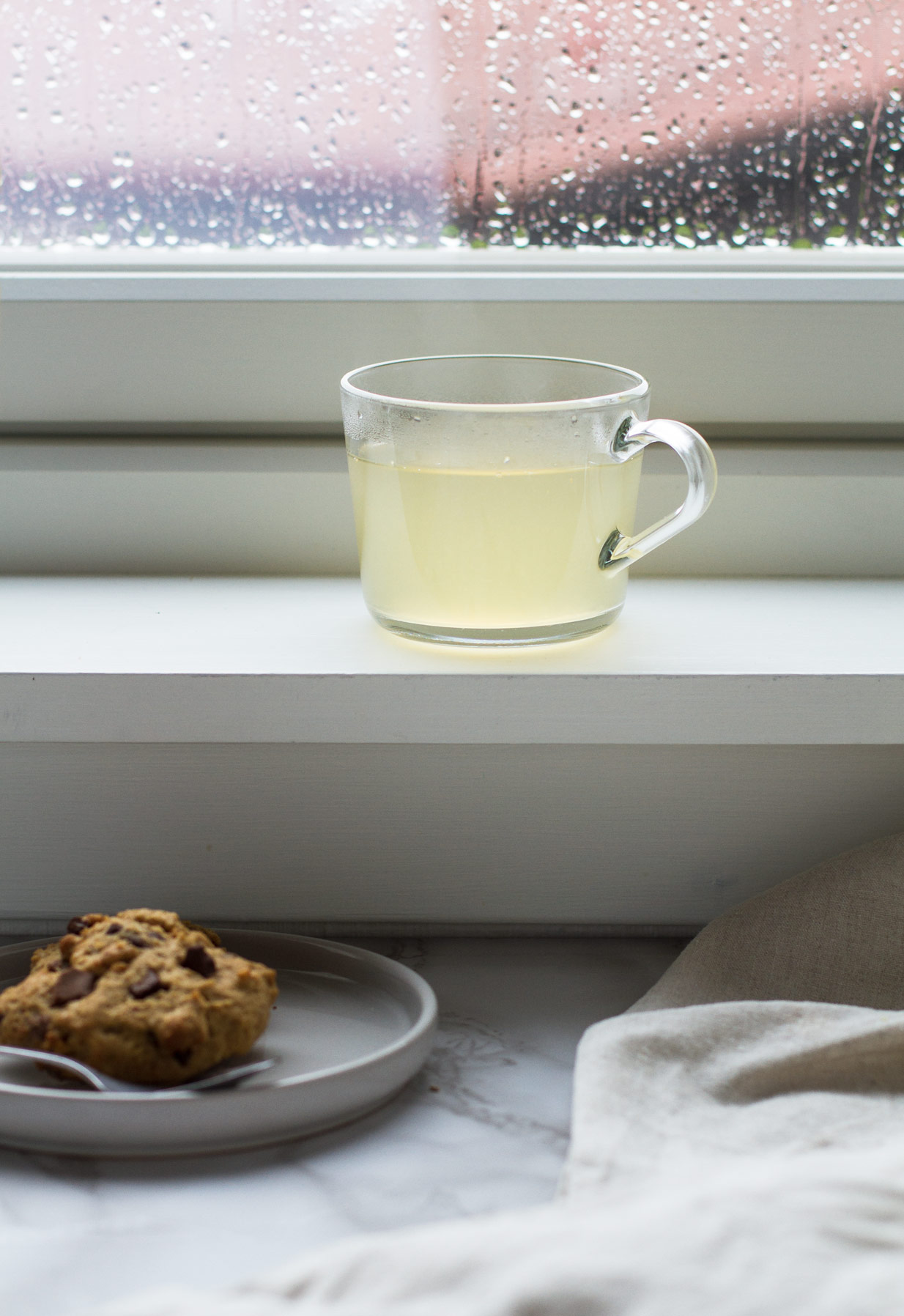 Clear mug with pale yellow drink in the window frame. Cookies on the table.