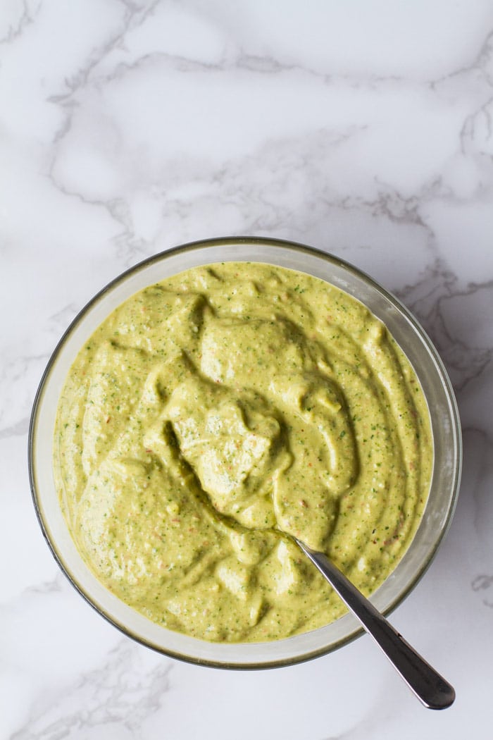 Green avocado sauce in a clear bowl and a spoon. White marble background.