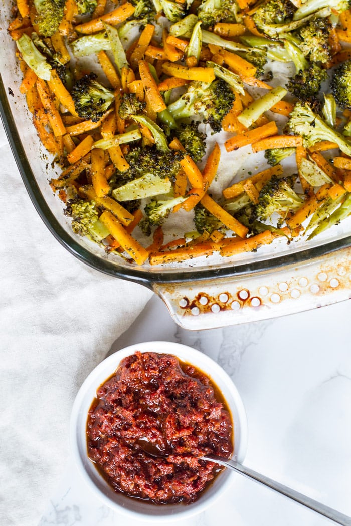 A clear baking dish with roasted broccoli and carrots. A white ramekin with sun dried tomato paste and a spoon. 