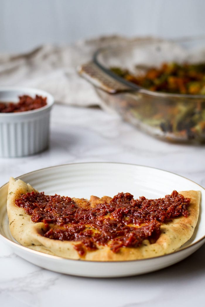 A naan spread with sun dried tomato paste on a white plate. Ramekin with sun dried tomatoes in the background blurred out.