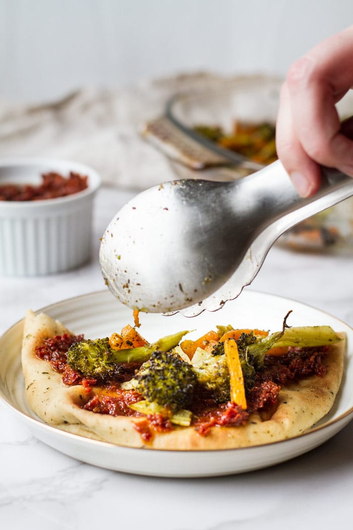 Naan with sun-dried tomatoes and adding roasted broccoli and carrots with a metal tong.