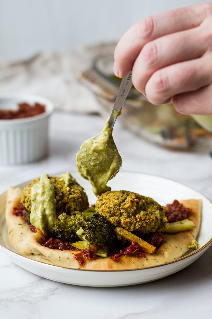 A naan with sun dreid tomatoes, roasted vegetables, baked falafel and drizzling avocado sauce with a spoon.