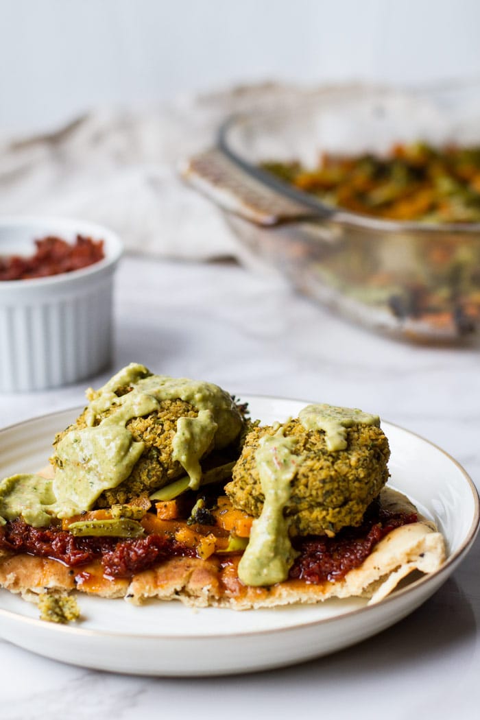 A naan with sun-dried tomatoes paste, roasted vegetables baked falafel patties and avocado sauce on a white plate and white marble background.