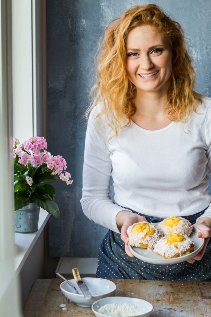 Girl with red hair and white shirt holding a beige plate with three vanilla custard and coconut sweet buns. Pink flower in the back.