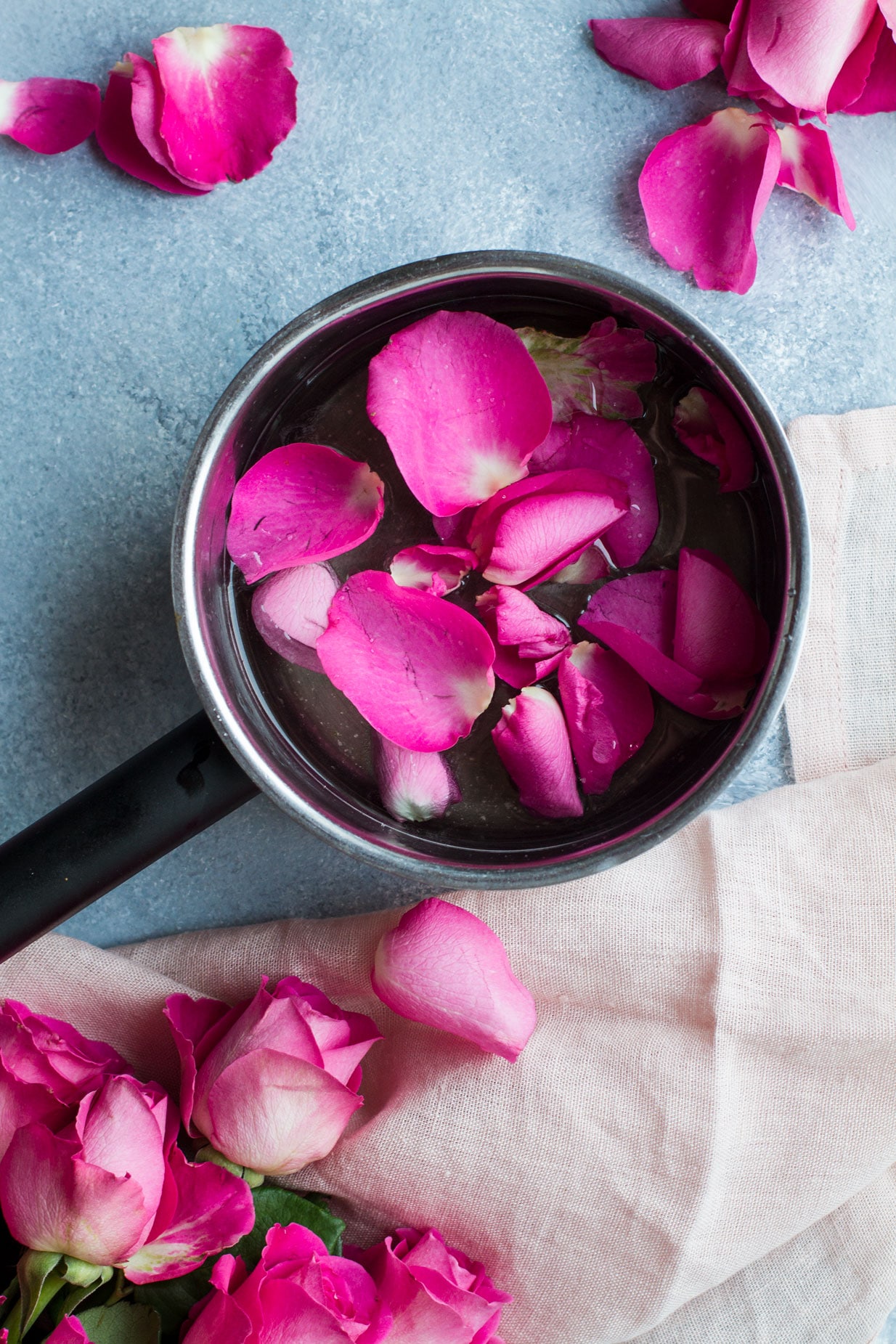 Bird's eye view of a saucepan with pink rose petals. Roses in the bottom left corner, blue background and pink cloth.