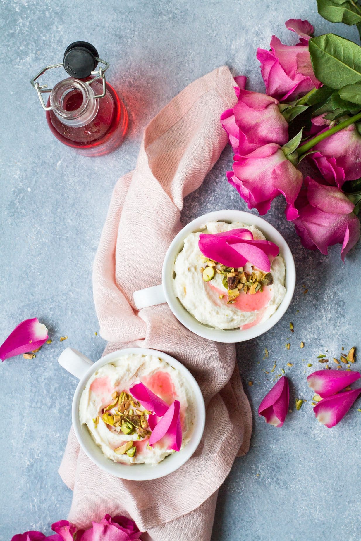 Bird's eye view of two white cups with mousse, chopped pistachios and pink rose petals. Rose petals around the blue table, pink cloth.