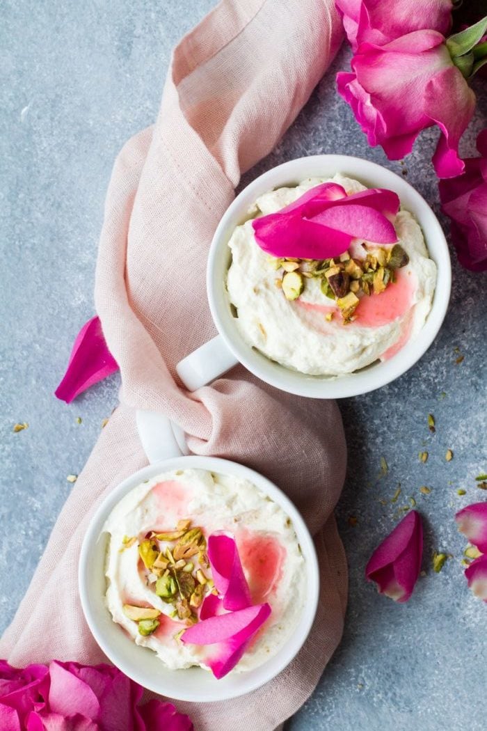 Bird's eye view of two white cups with white chocolate mousse, chopped pistachios and pink rose petals. Rose petals around the blue table, pink cloth.