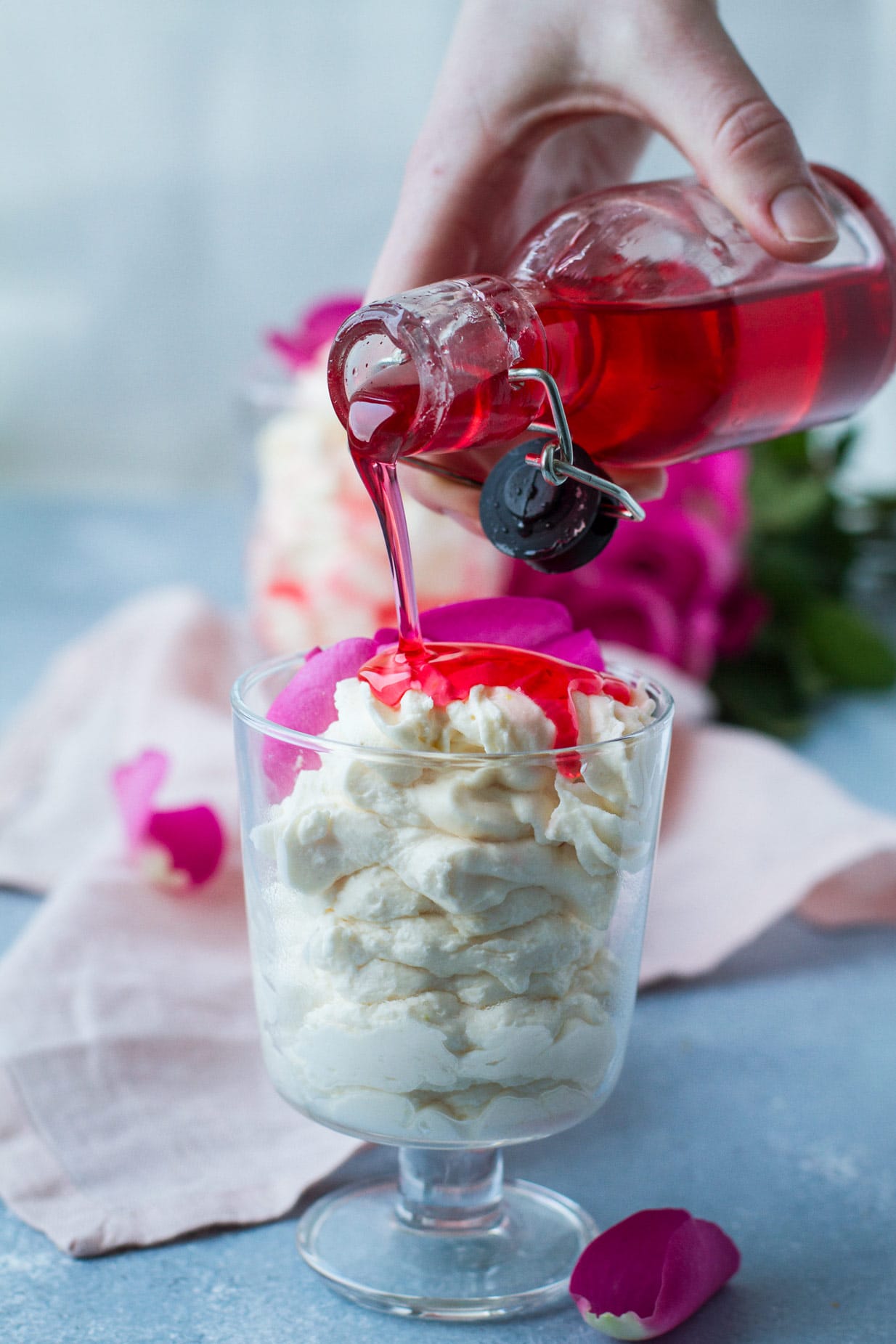 A glass with white mousse and a glass of red rose syrup pouring over the white mousse. Blue table and pink cloth, blurred pink roses in the background. 