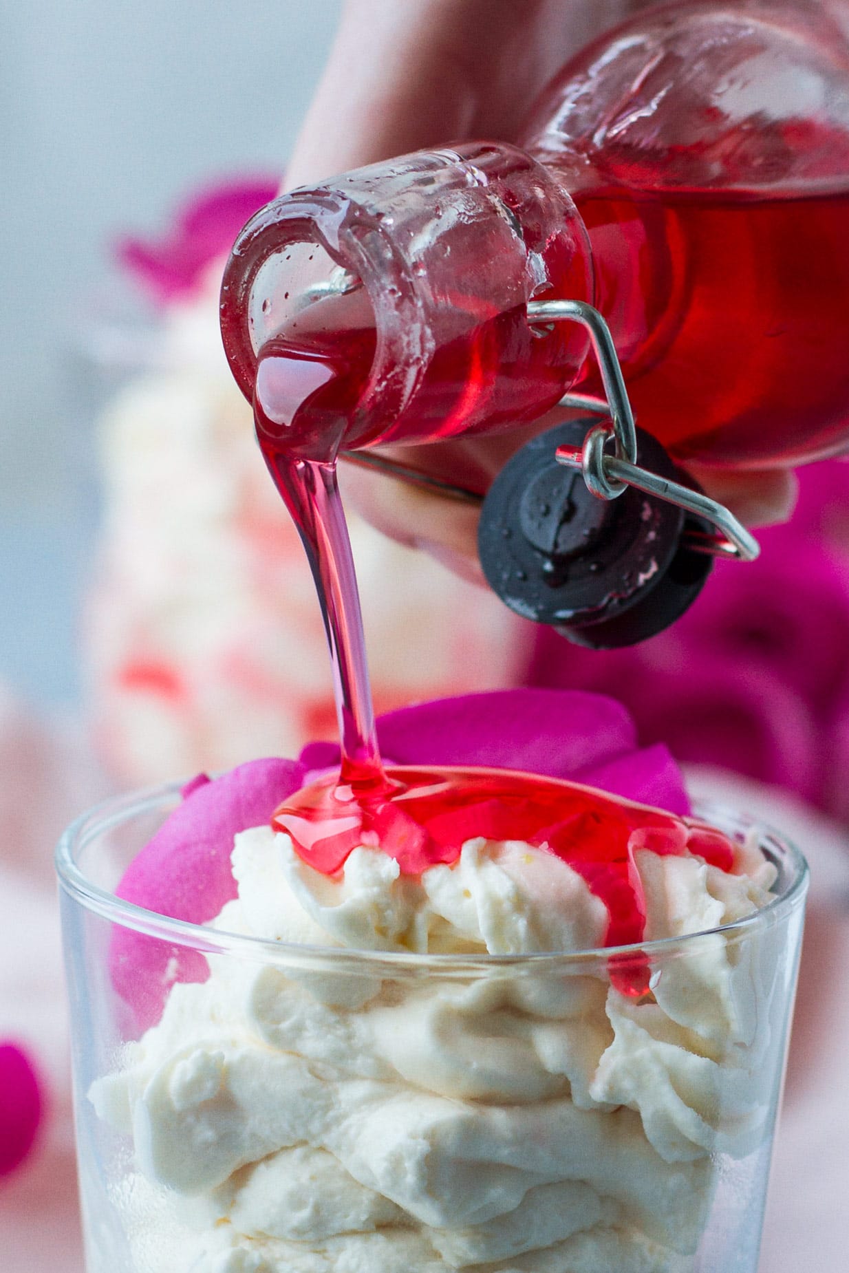 Closeup. A glass with white mousse and a glass of red rose syrup pouring over the white mousse. Blue table and pink cloth, blurred pink roses in the background. 
