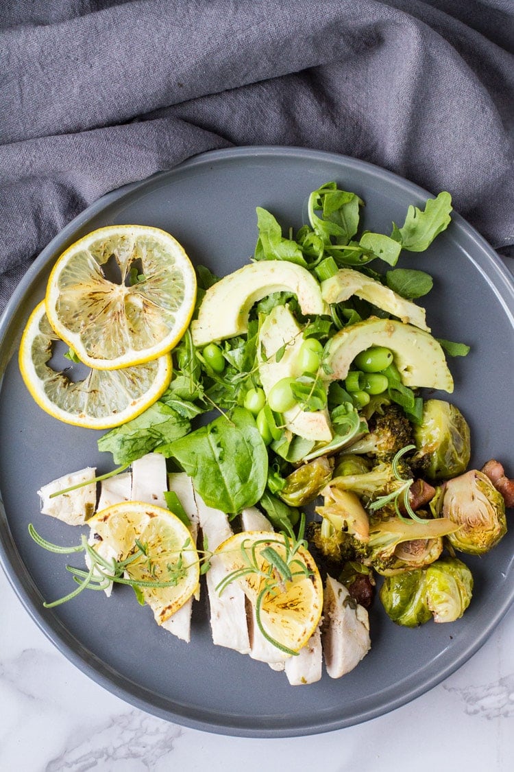 Flat lay of baked lemon chicken with green spring salad and lemon slices, dark grey plate.