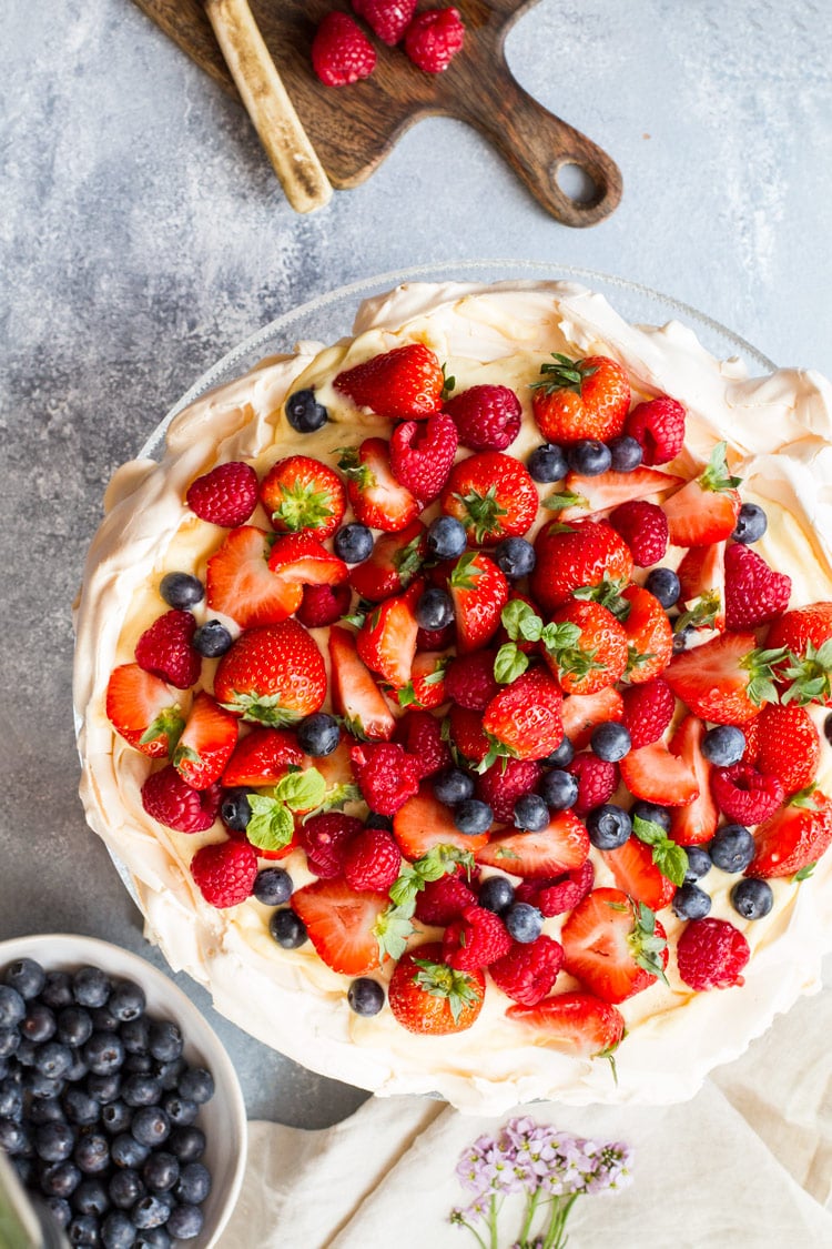 Flatlay of this pavlova recipe with fresh berries. A bowl of blueberries on the side.