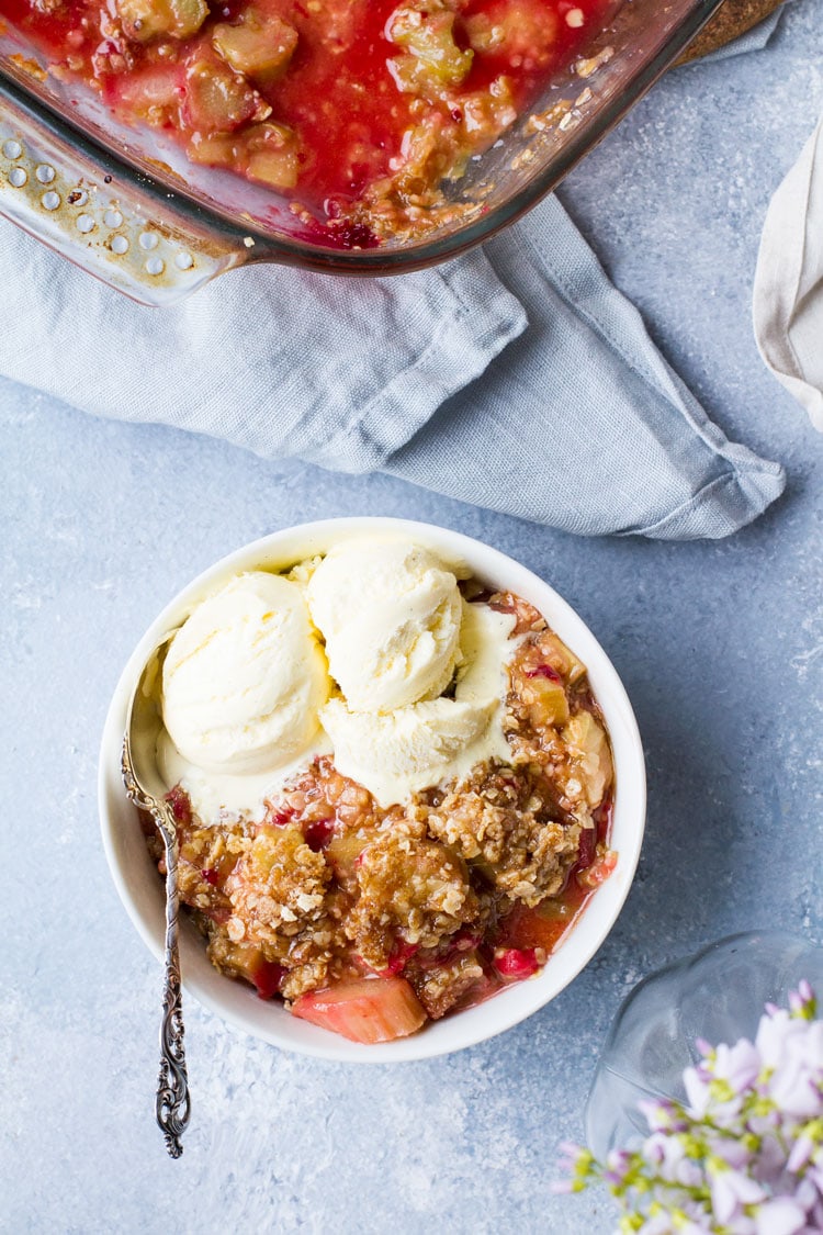 Flatlay of white bowl with raspberry rhubarb crisp and vanilla ice cream. Blue background.