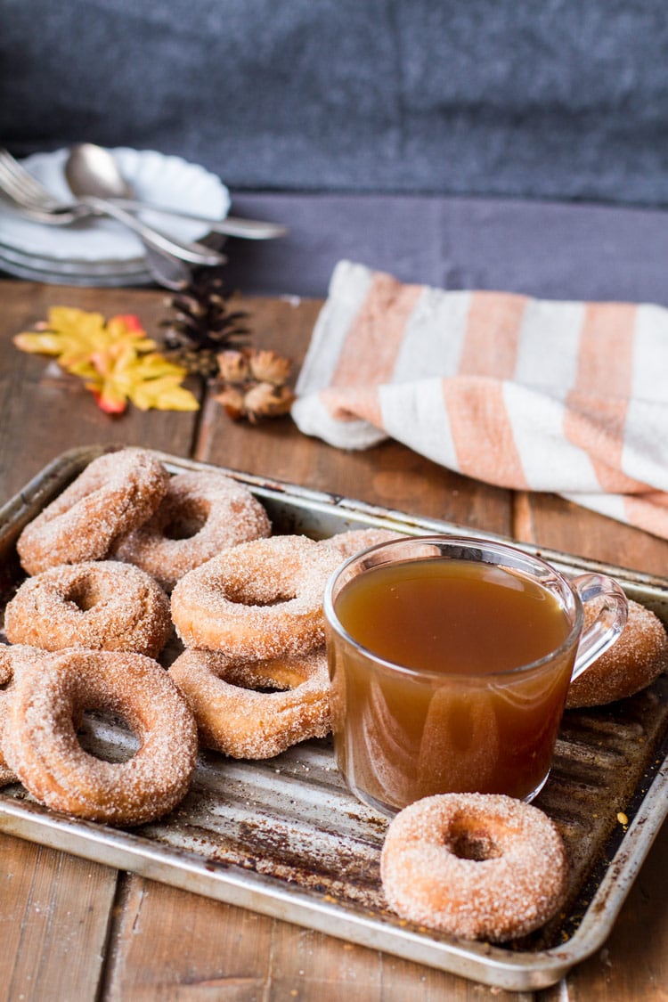 A metal pan with apple cider donuts and apple cider in a glass cup. Orange striped towel in the back.