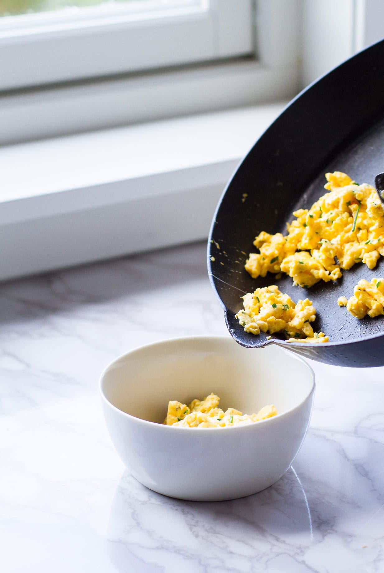Pouring scrambled eggs from skillet to a white bowl.