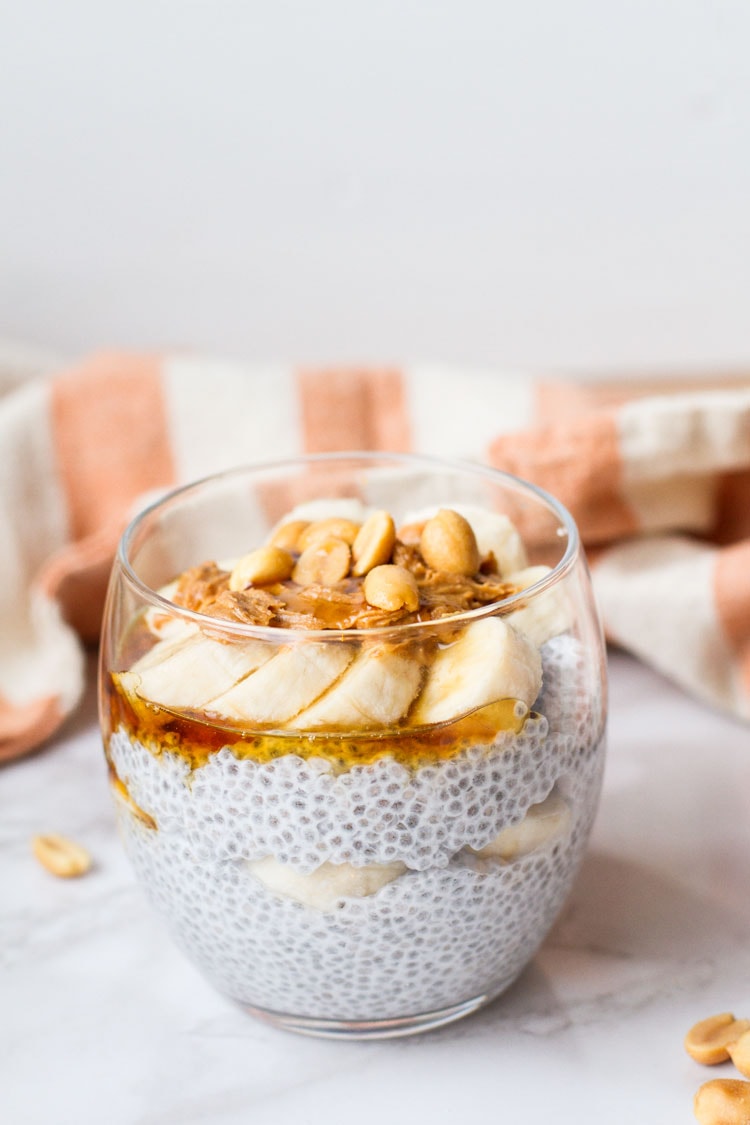 Round glass with chia pudding topped with sliced banana, peanut butter and peanuts. White background.