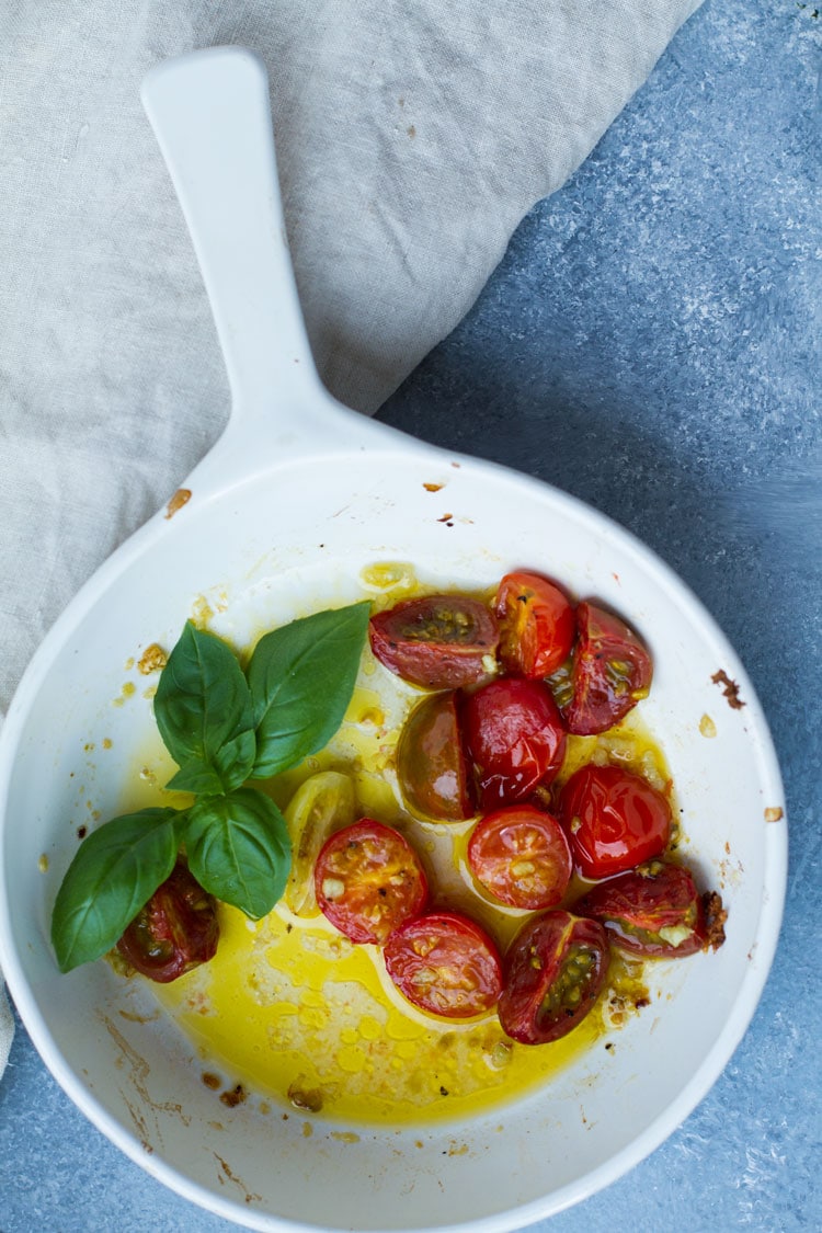 White dish with roasted cherry tomatoes in garlic and olive oil. Garnished with fresh basil.