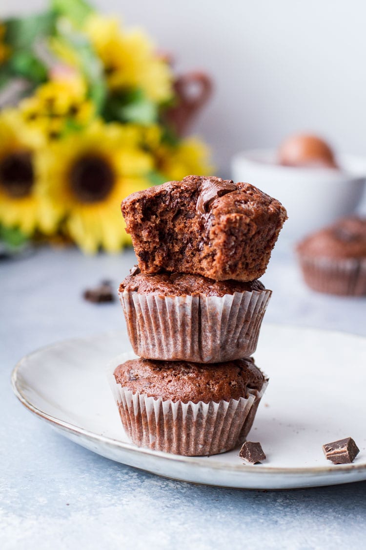 Three chocolate banana muffins on top of each other, top one is taken a bite out of. Sunflowers in the background.
