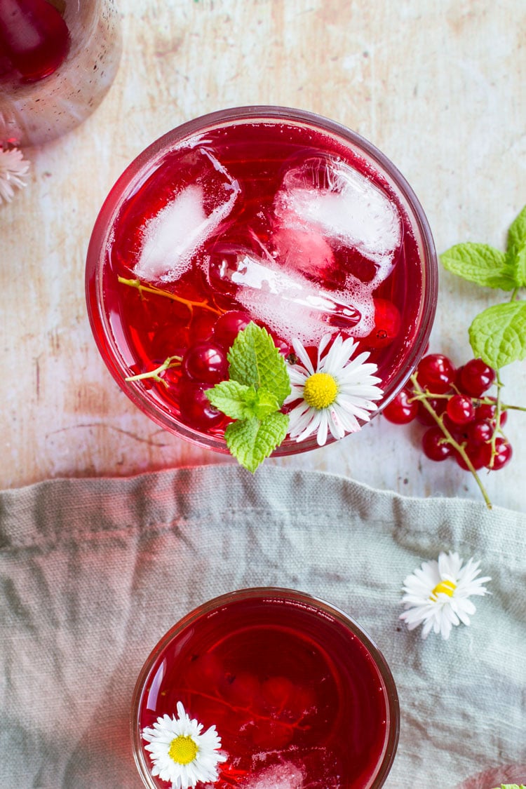 Glass with red drink and ice cubes, garnished with flower, mint leaves and red currants. Flatlay.
