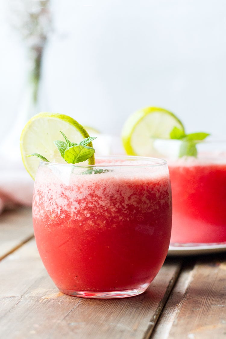 Straight on view of two round glasses with watermelon drink and lime wedges.