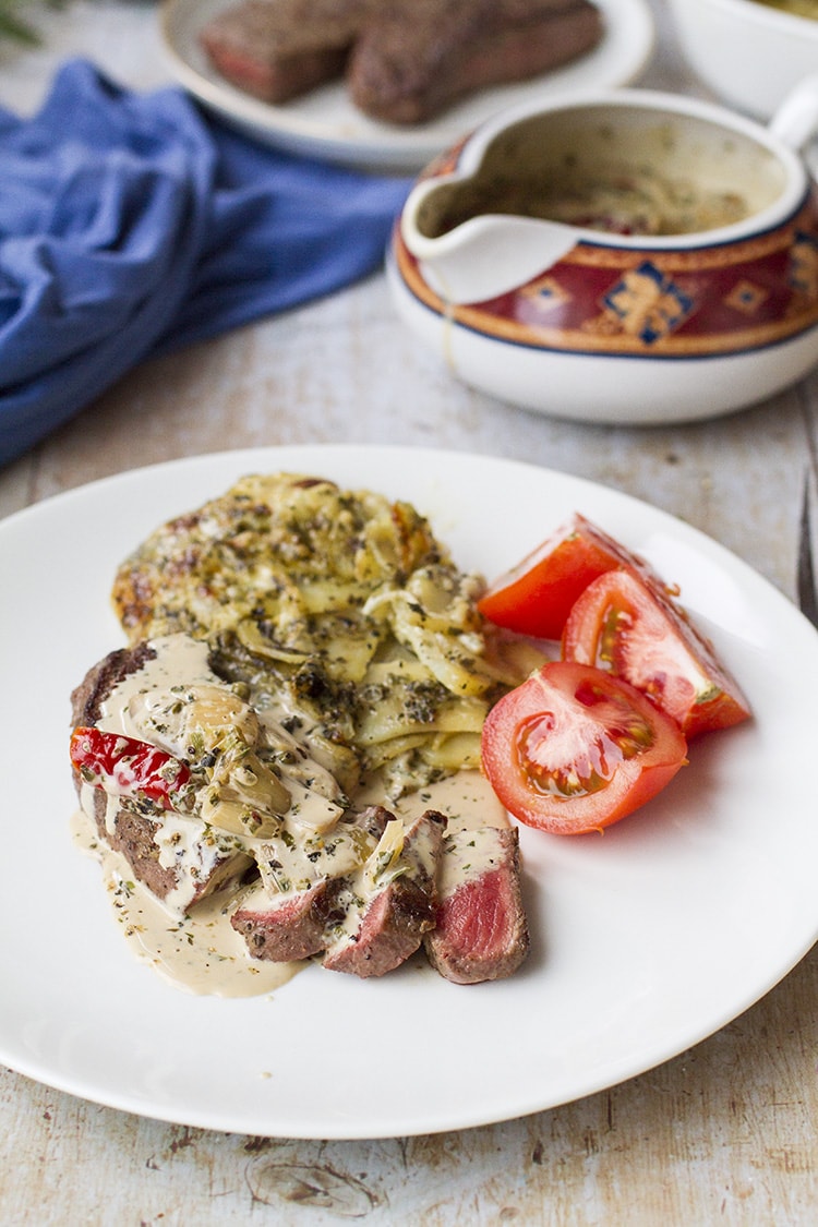 Complete dish on a white plate with the sauce, steak, tomatoes and scalloped potatoes.
