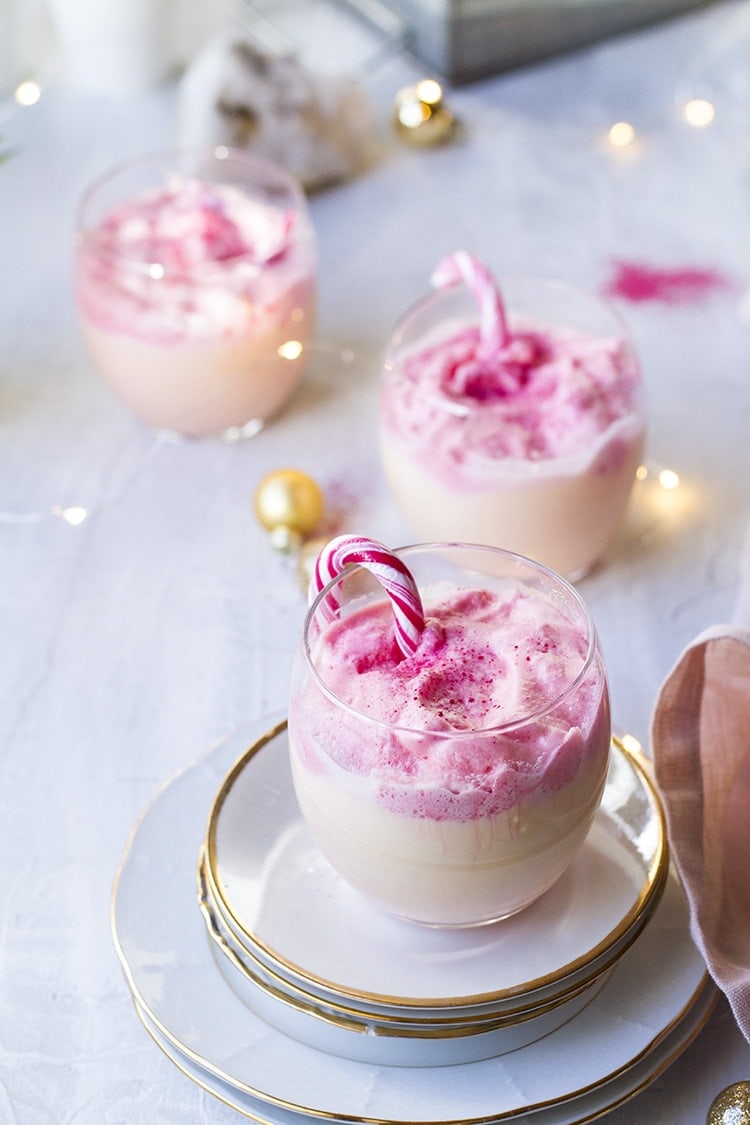 Three glasses with eggnog and pink dalgona, garnished with candy canes.
